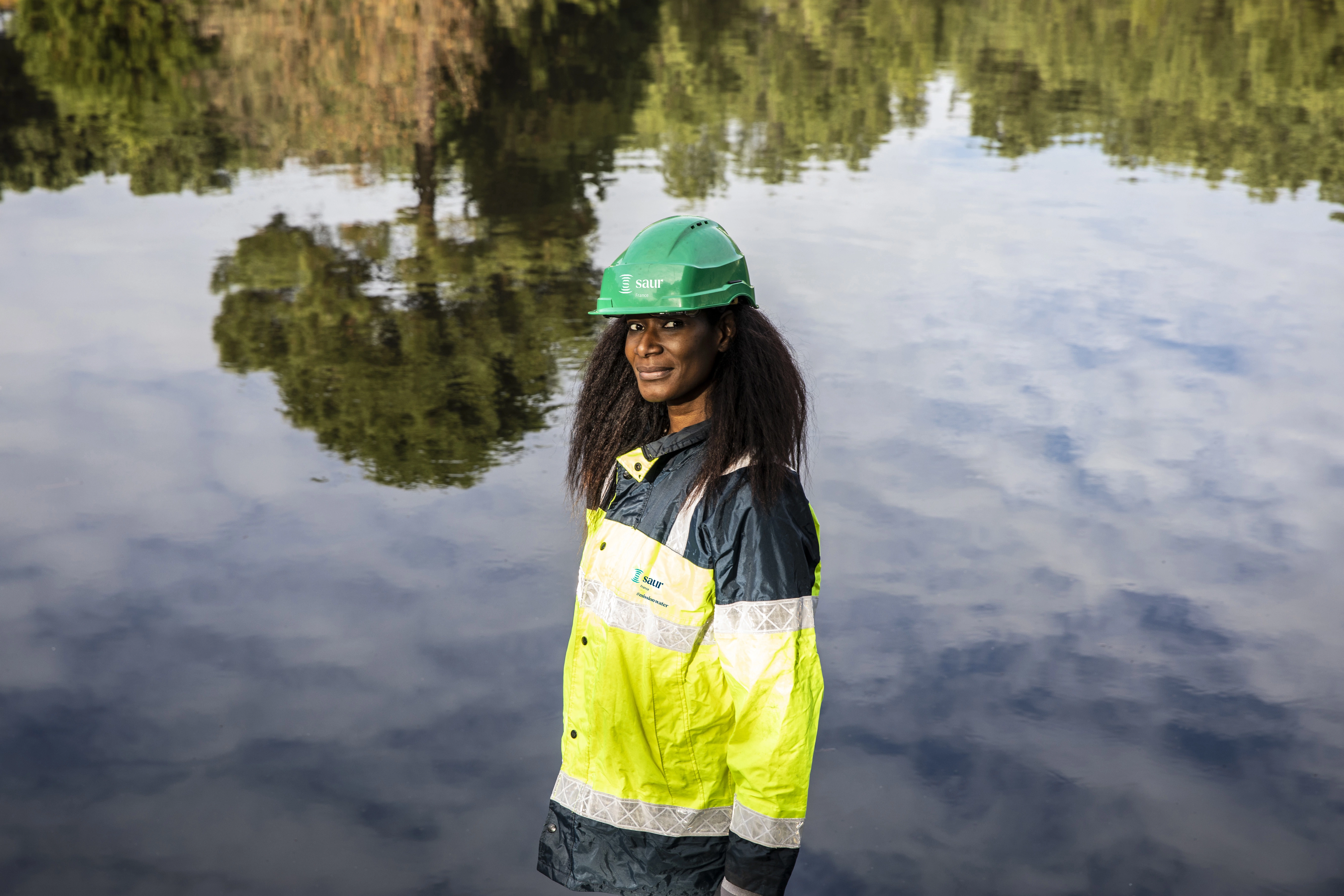 A woman working for Saur France, wearing an equipment, standing next to water, smiling at the camera
